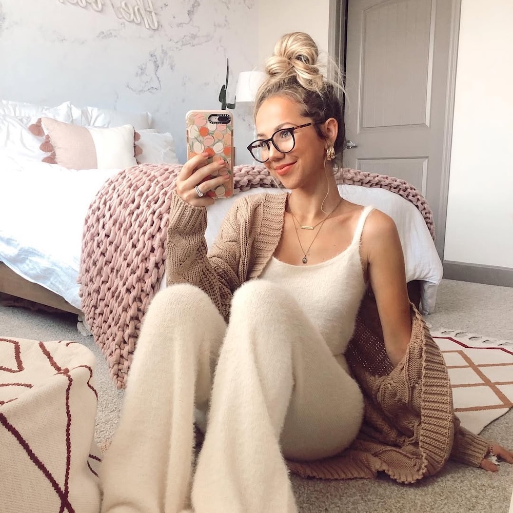 A woman wearing cozy loungewear and glasses sits on a bedroom floor, smiling and taking a mirror selfie with her phone. The room has a neatly made bed, soft lighting, and neutral, textured decor.