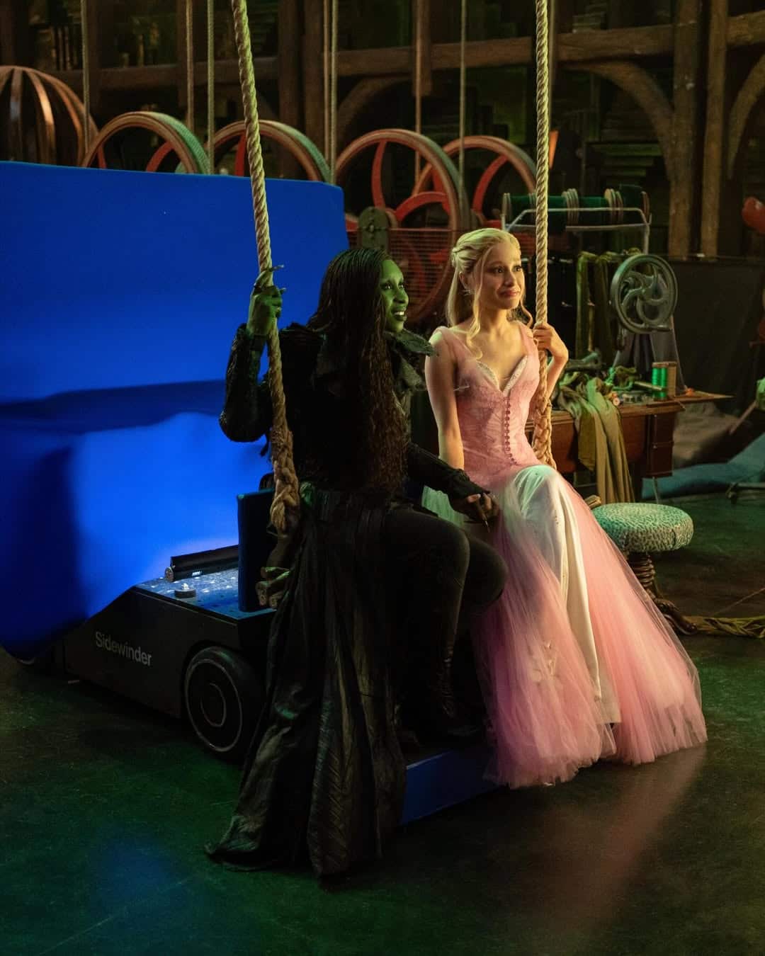 Two actors in costume sit on a swing; one is painted green and wears a long black coat, the other wears a pink gown and tiara. Behind them are stage equipment and a blue screen, hinting at a Wicked Part 2 production set before it hits theaters.