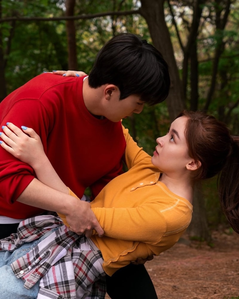 A young man in a red sweatshirt dips a surprised young woman in a yellow top while standing in a wooded area, both gazing into each other's eyes, as if they're about to choose love like Kitty in XO Kitty Season 3.
