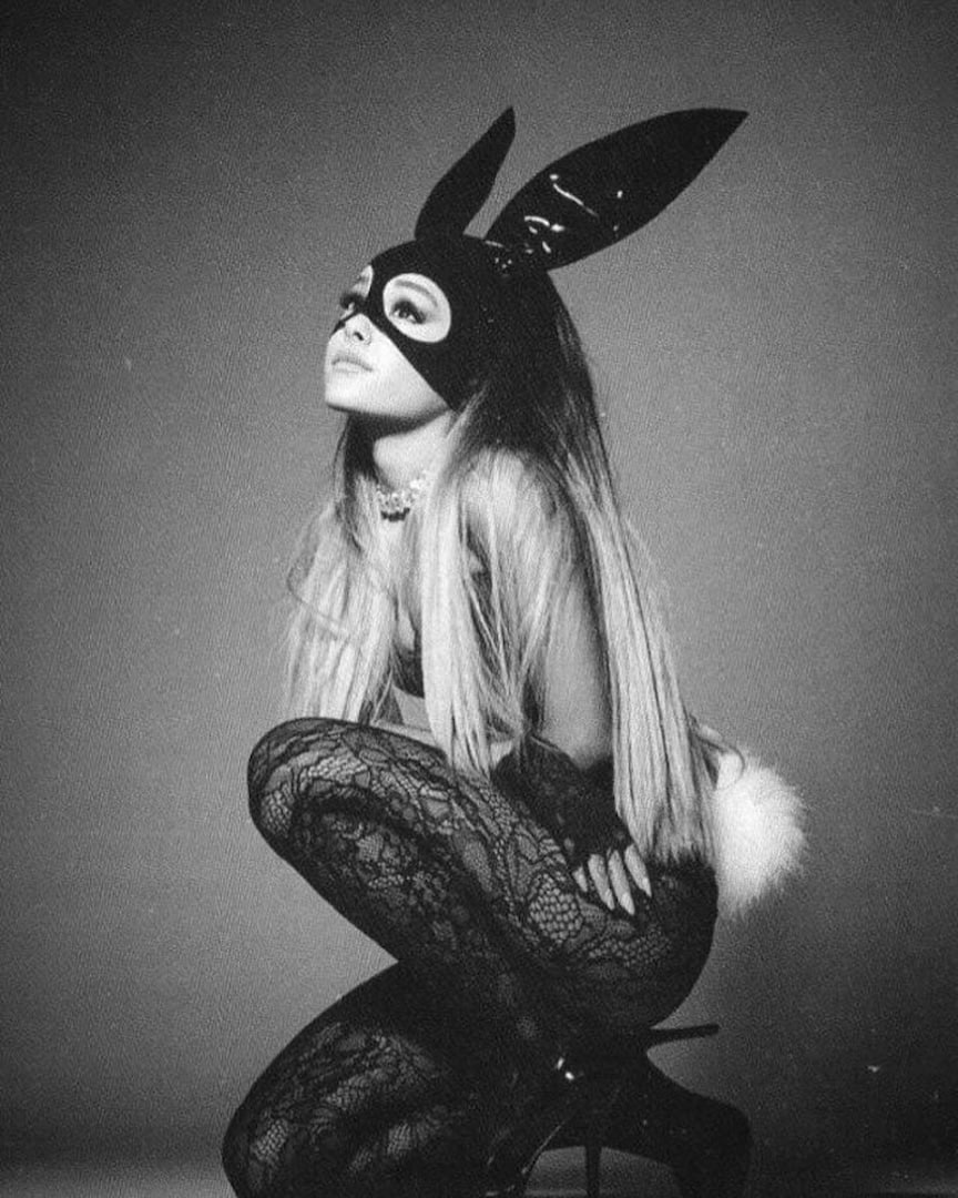 A woman in a black lace bodysuit, bunny ears mask, and a white fluffy tail poses in profile against a plain background. She is crouched on a stool, looking upward, and the image is in black and white.