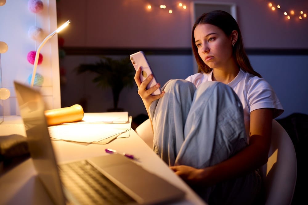 A teenage girl sits at a desk with a laptop and papers, looking at her phone with a serious expression. She hugs her knees to her chest in a dimly lit room decorated with string lights, worrying about communication issues with her boyfriend not responding.
