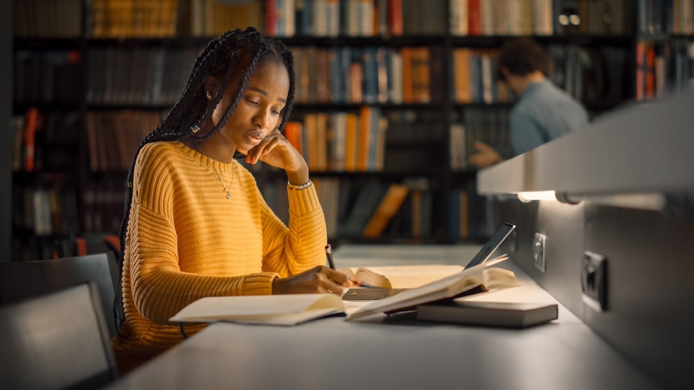 A young woman wearing a yellow sweater studies at a desk in a library, surrounded by open books, as she prepares to write papers for school. Shelves of books and another person appear in the background.