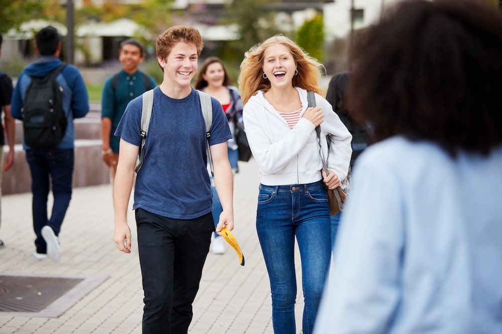 Two teenagers, a boy and a girl, are walking outside on a paved path, smiling and talking. Both wear casual clothes and backpacks, perhaps sharing dating advice or relationship tips as other students pass by in the background.