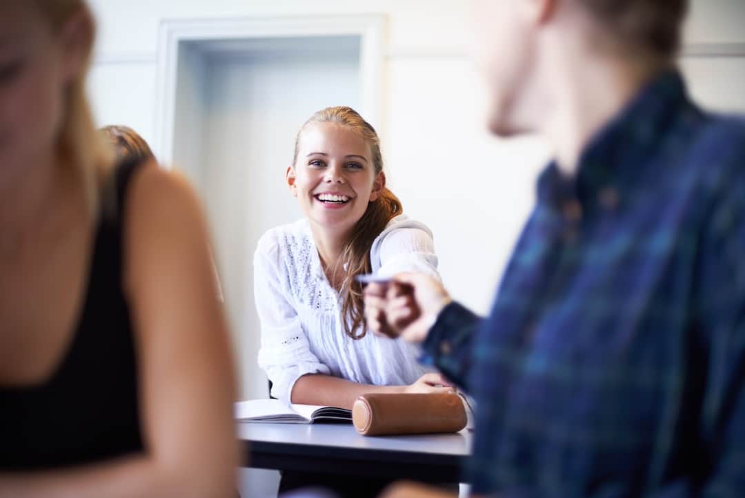 A young woman in a white shirt smiles while sitting at a desk in a classroom, with an open notebook and pencil case in front of her, as someone in the foreground offers relationship advice and gestures animatedly.