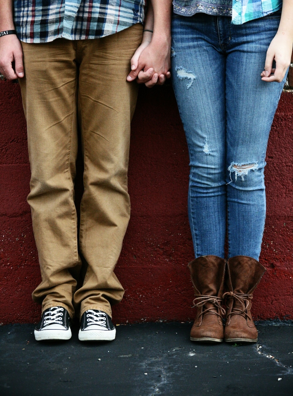 Two people dating stand side by side, holding hands. Only their legs and torsos are visible—one in khaki pants and black sneakers, the other in ripped blue jeans and brown boots—both against a red wall, perhaps hiding from strict parents.