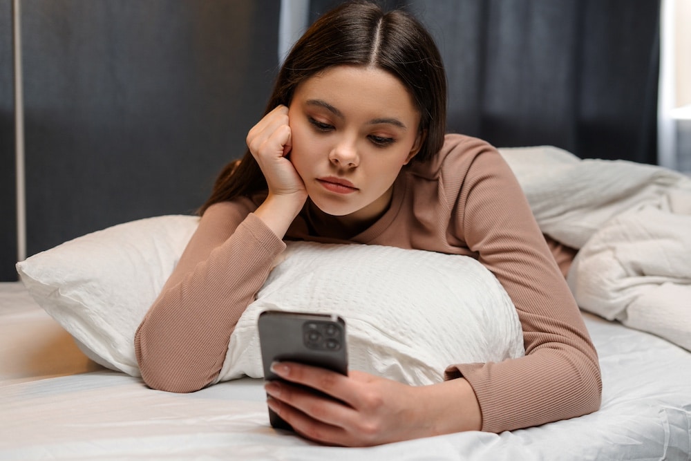 A young woman with long brown hair lies on a bed, propping her head on her hand, looking at her smartphone with a thoughtful expression—perhaps reflecting on dating regret or seeking relationship advice. She wears a light brown long-sleeve top.