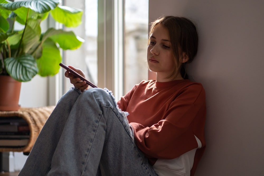 Carol, dressed in a rust-colored sweatshirt and jeans, sits on the floor indoors, leaning against a wall as she looks thoughtfully at her phone. Sunlight streams through a window, while a potted plant adds calm to her quiet moment of feelings.
