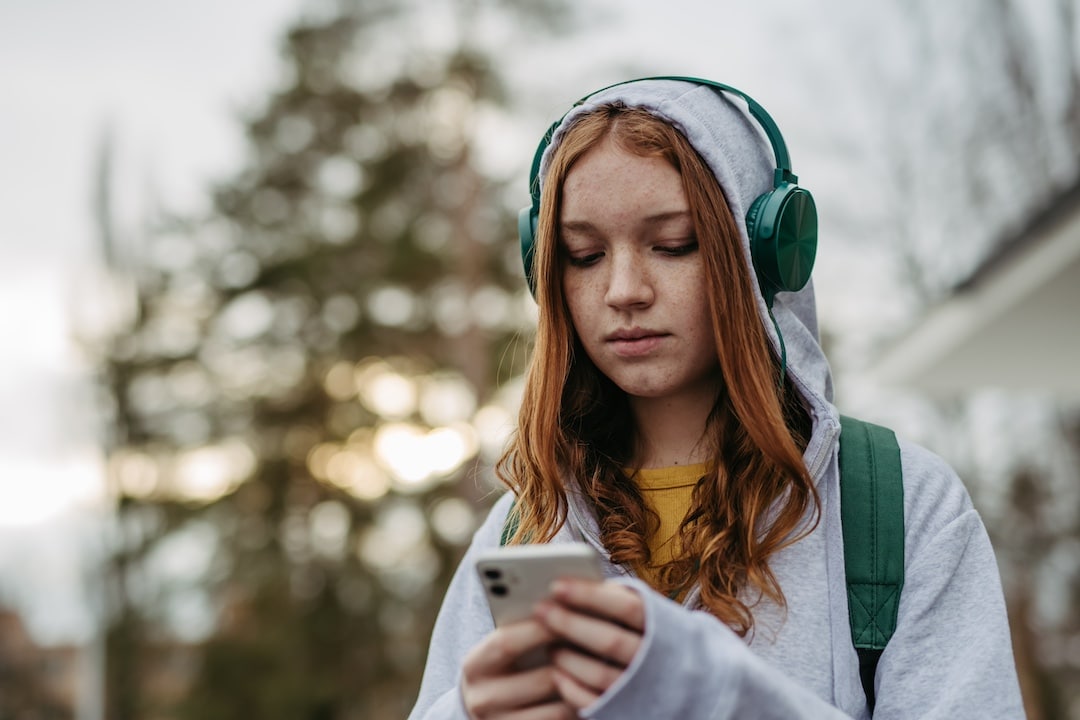 A teenage girl with long red hair, wearing a gray hoodie and green headphones, looks at her smartphone outdoors—perhaps texting her crush or seeking relationship advice amid trees and a blurred building in the background.
