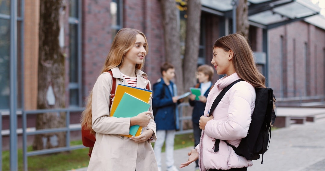 Two teenage girls with backpacks talk and smile outside a school building, holding notebooks, as one asks her friend for advice because she’s confused about feelings. In the background, two boys also talk near trees and brick walls.