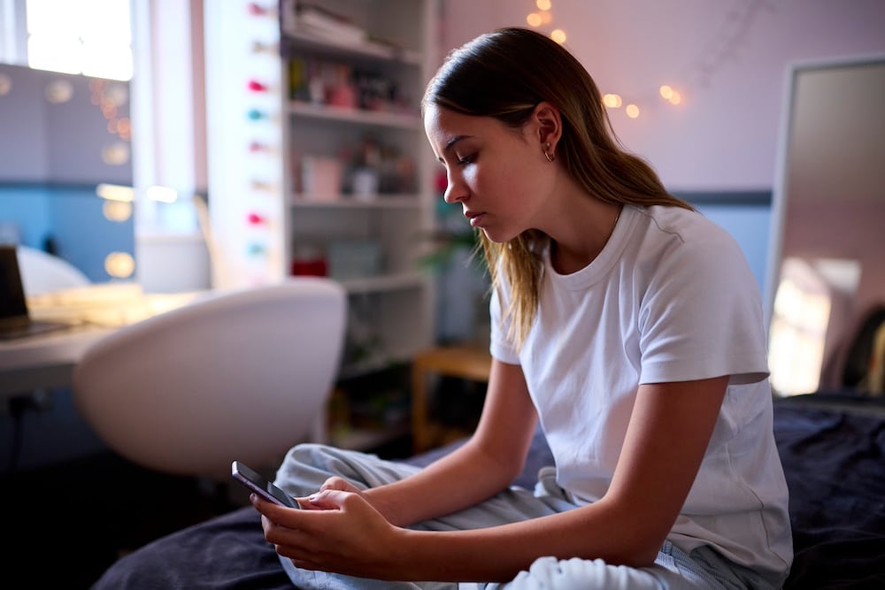 Teen girl sitting on her bed in a cozy, softly lit bedroom, looking down at her smartphone with a thoughtful, slightly stressed expression. String lights decorate the wall and a white chair sits by a desk in the background.