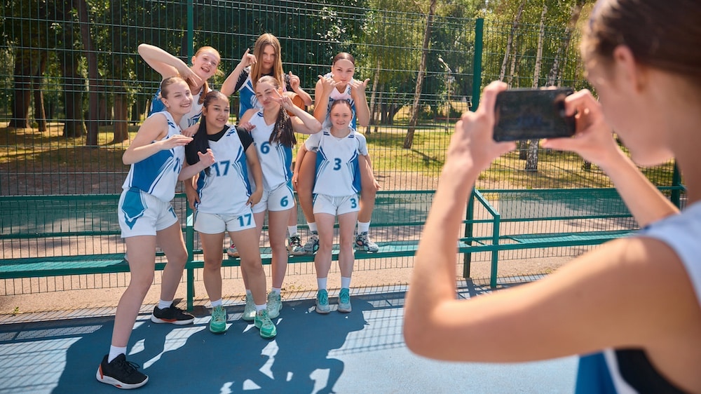 A group of teenage girls in basketball uniforms pose together and smile while Carol stops to take their photo outdoors on a sunny day. A metal fence and trees are in the background.