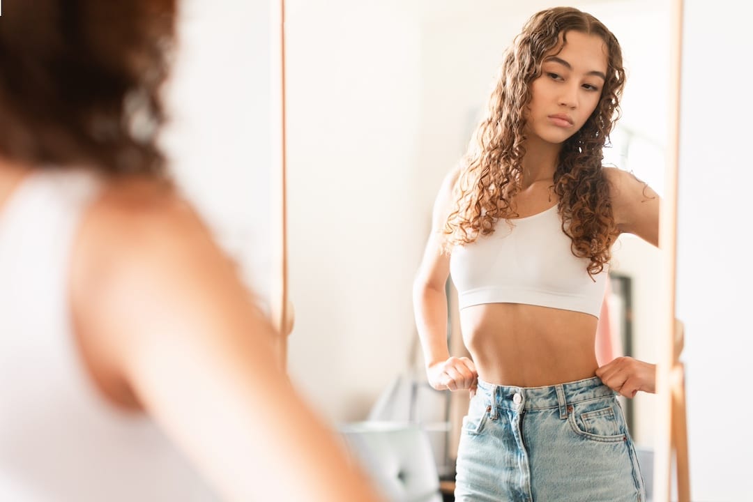 A young woman with long curly hair wearing a white crop top and jeans stands in front of a mirror, looking at her reflection with a thoughtful, self-conscious expression and her hands on her hips, noticing her small boobs.