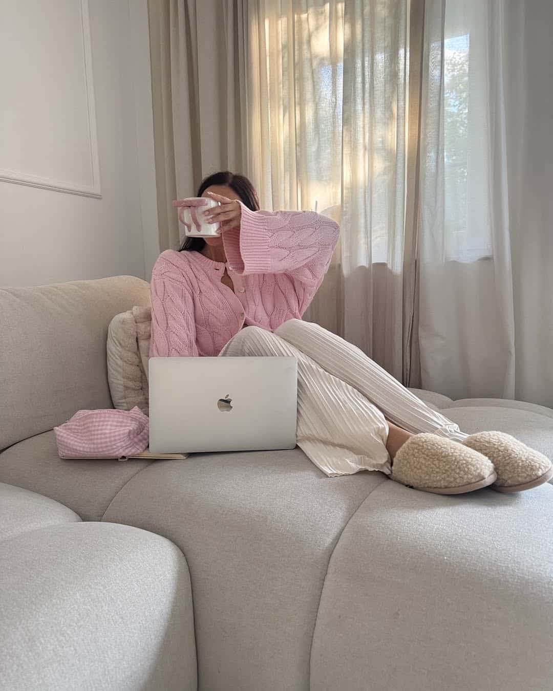 A person in cozy loungewear sits on a light gray couch with a laptop on their lap, holding a mug in front of their face—unwinding as part of their after-school routine. Soft natural light streams through sheer curtains in the background.