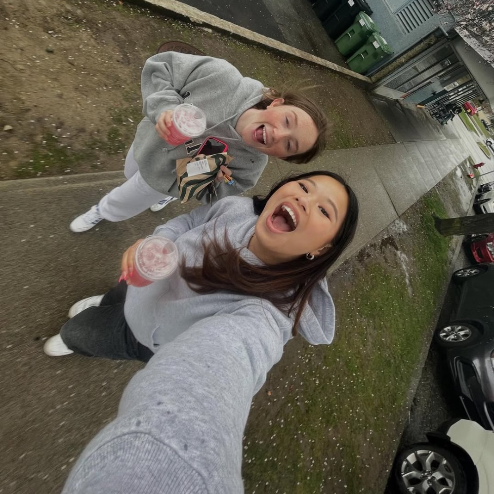 Two young women stand outdoors on a sidewalk, smiling and holding drinks. One is holding a coffee carrier. They are both wearing casual, comfortable clothing and appear happy, posing for a selfie on a cloudy day.