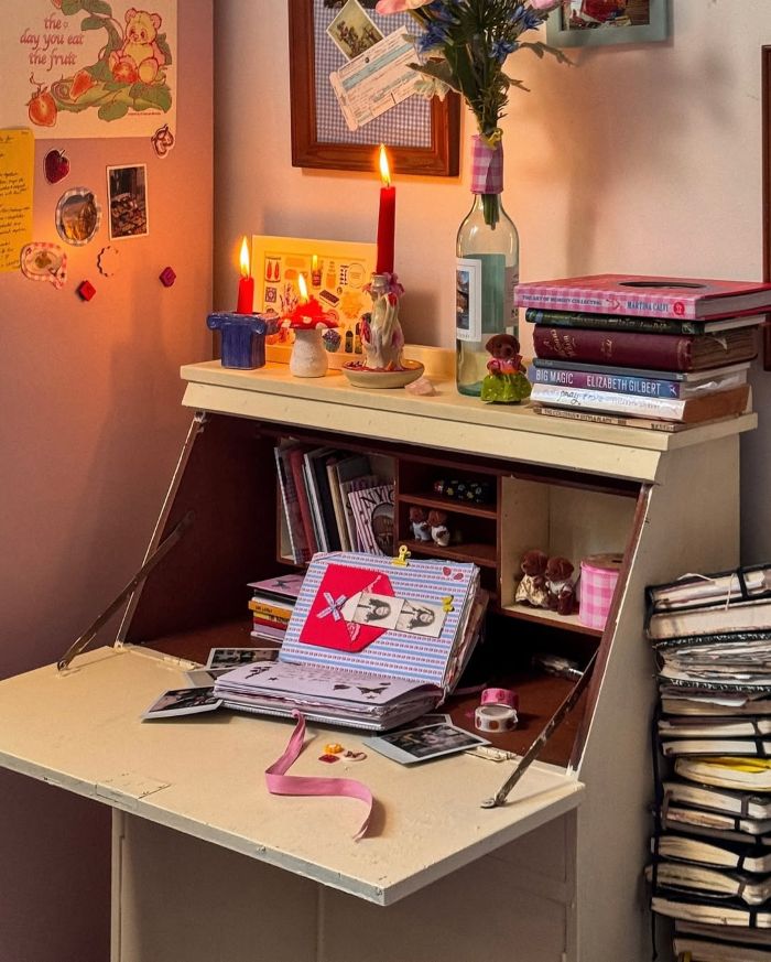A cozy desk with an open writing surface displays books, photos, stationery, and pink ribbons—perfect for quick home decor. Above, candles burn in wine bottles with flowers, while notes and pictures on the wall create a warm, creative atmosphere.