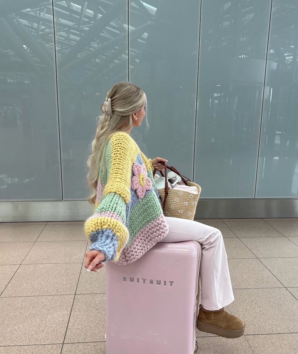 A woman with long blonde hair sits on a pink SUITSUIT suitcase, wearing a colorful flowered sweater, white pants, and brown boots, holding a woven bag and her latest book to read as she awaits her dream travel in the airport terminal.