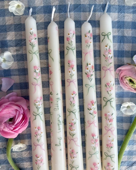 Five white taper candles with hand-painted pink flowers and green leaves are arranged on a blue and white checkered cloth, surrounded by pink and white flowers and flower petals.