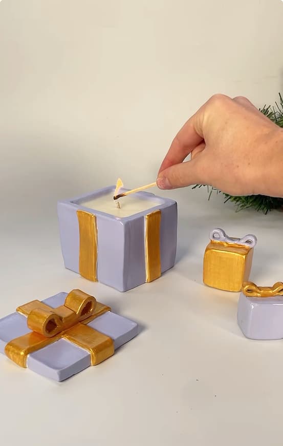A hand lights a candle shaped like a gift box with a match. The candle and two small gift-shaped containers are purple with gold ribbons, and a matching gift-shaped tray is nearby.