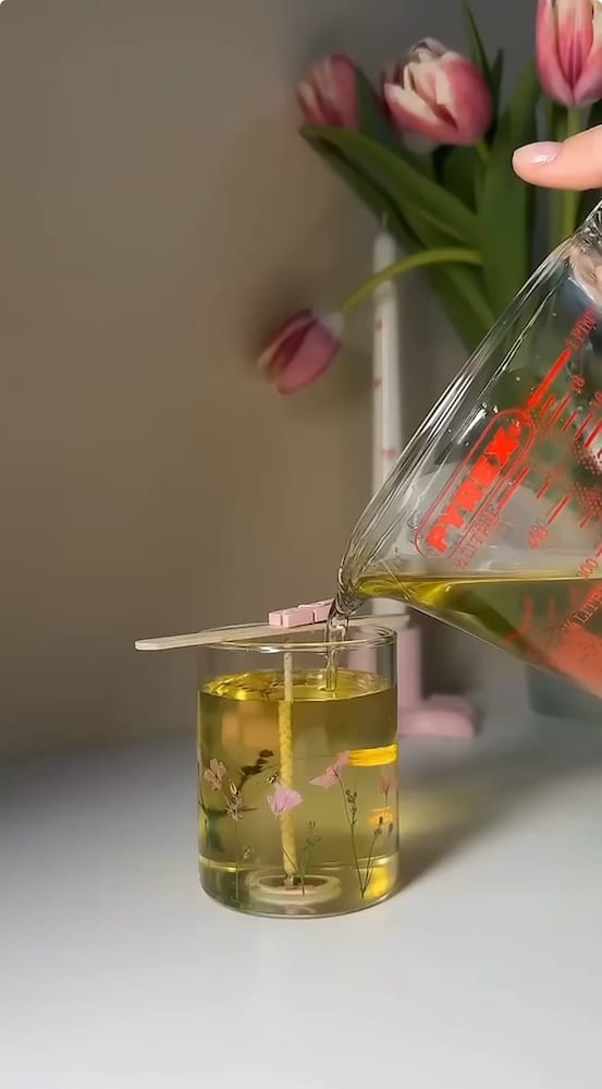 A hand pours melted wax from a glass measuring cup into a clear, decorated candle jar with a centered wick, with pink tulips and a white surface in the background.