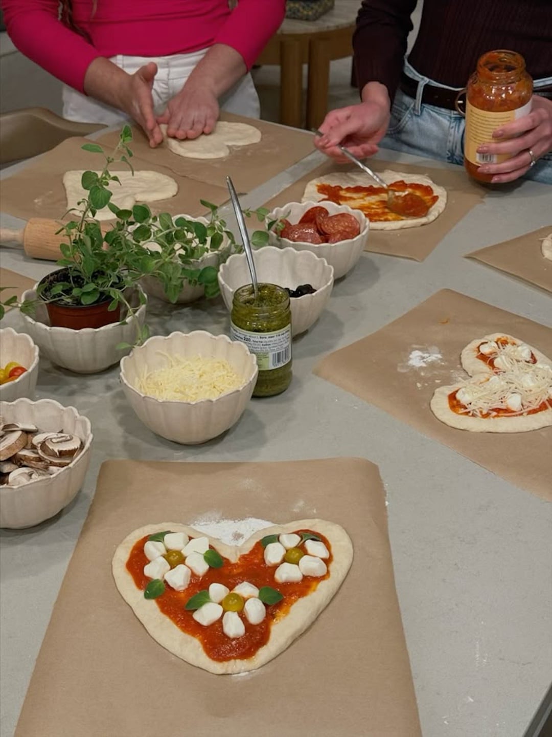 Two people enjoy a DIY pizza night, making heart-shaped pizzas topped with sauce, mozzarella, and basil. Bowls of cheese, mushrooms, olives, and herbs surround them—perfect for anyone wanting to host the best homemade pizza party.
