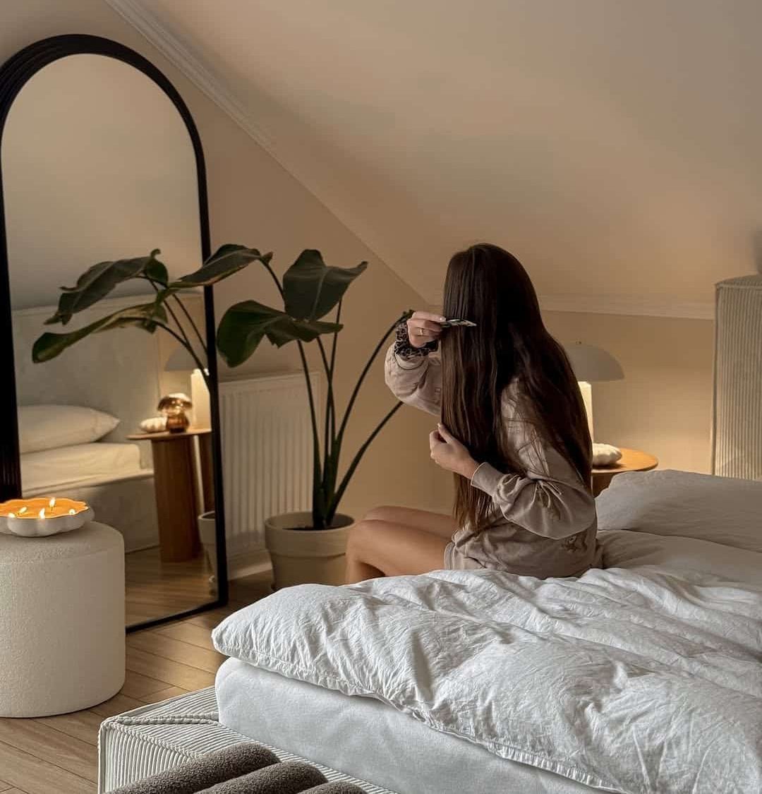 A woman sits on a neatly made bed in a cozy, softly lit bedroom, brushing her long hair. A large mirror, green plant, and a lit candle on a side table add warmth to the serene space.
