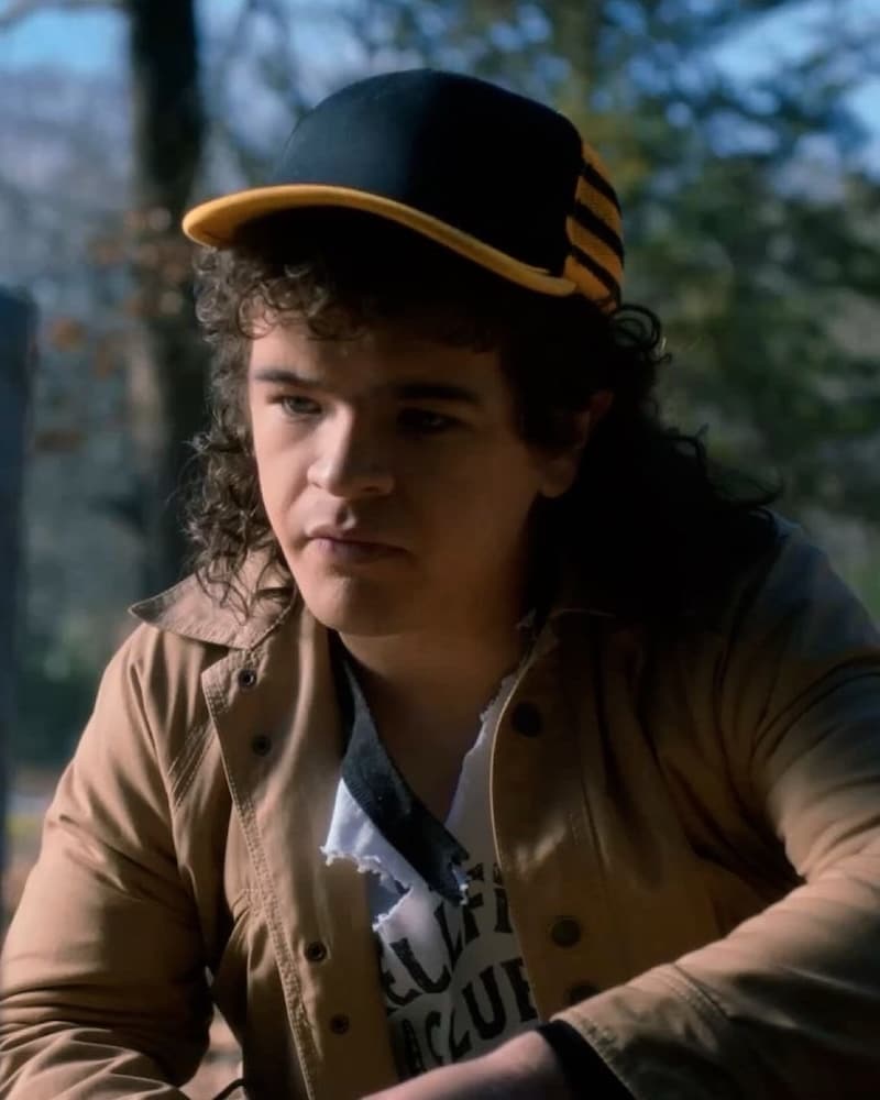 A young person with curly brown hair wears a black and yellow cap, beige jacket, and white t-shirt with a torn collar, sitting outdoors with trees blurred in the background.