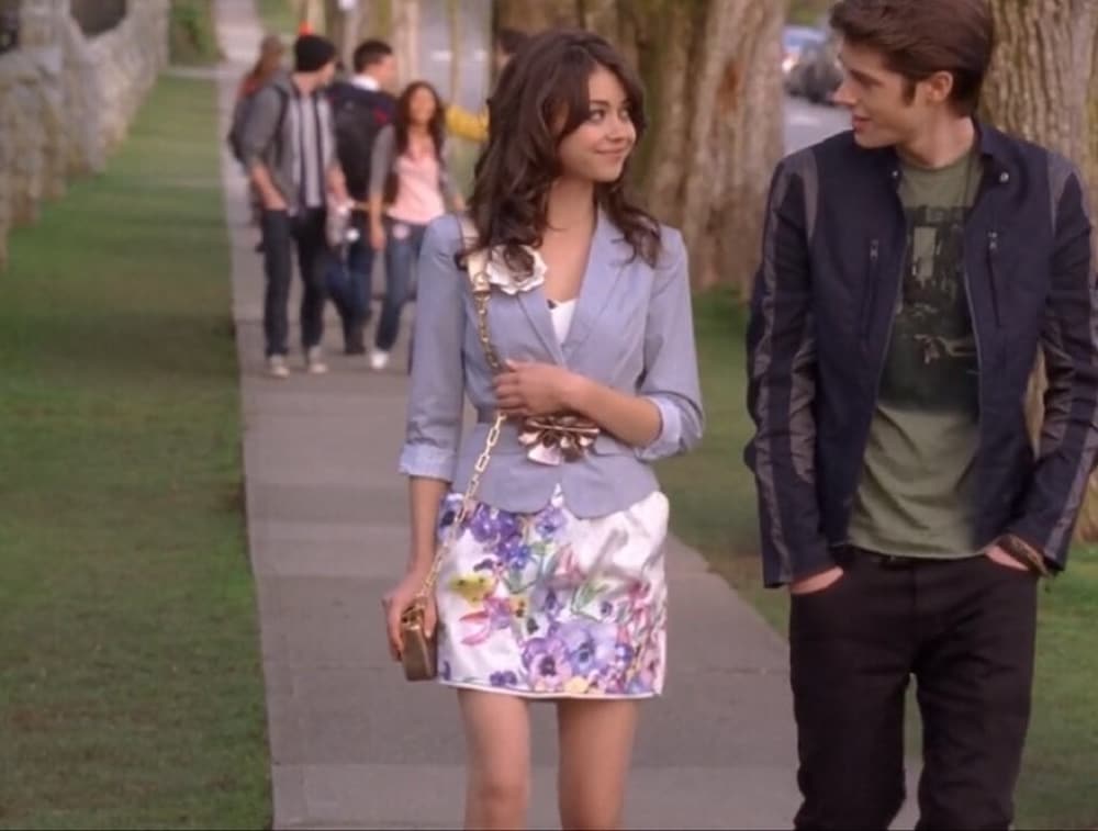 A young woman in a floral skirt and light blue blazer walks on a sidewalk, smiling at a young man in a black jacket—like a scene straight out of romantic comedies. Other teenagers stroll in the background along the tree-lined street.