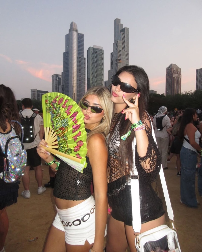 Two women in festival outfits and sunglasses pose together outdoors with city skyscrapers in the background. One holds a decorative fan; both wear black mesh tops and white bottoms. Other festival-goers are visible behind them.