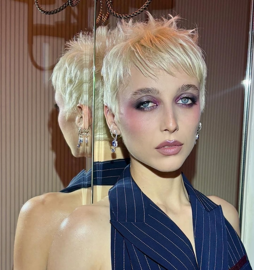 A person with short platinum blonde hair and dramatic dark eye makeup wears a navy pinstripe vest. They stand in front of a mirror, a retro telephone visible nearby, with serious facial expression and dangling earrings.