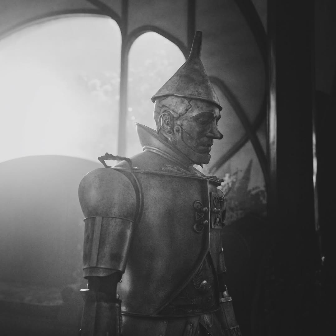 A person dressed as the Tin Man from The Wizard of Oz stands in profile indoors, illuminated by soft, dramatic light streaming through arched windows—evoking scenes from theaters or even Wicked Part 2. The image is in black and white.