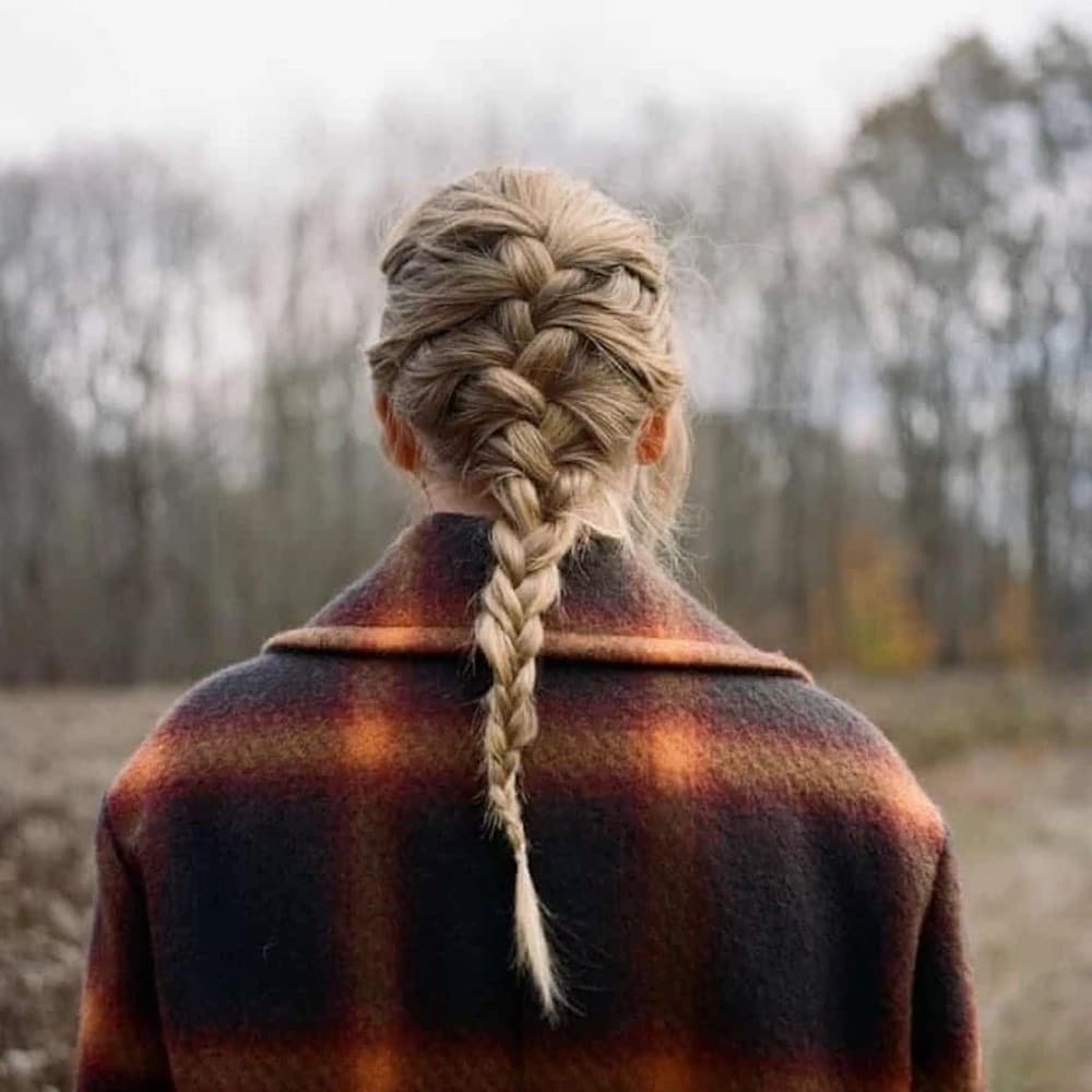 A person with blonde hair in a French braid stands outdoors, wearing a plaid coat—the scene evokes the moody vibe of a Taylor Swift album, with leafless trees and a cloudy sky hinting at a cool or autumn day.