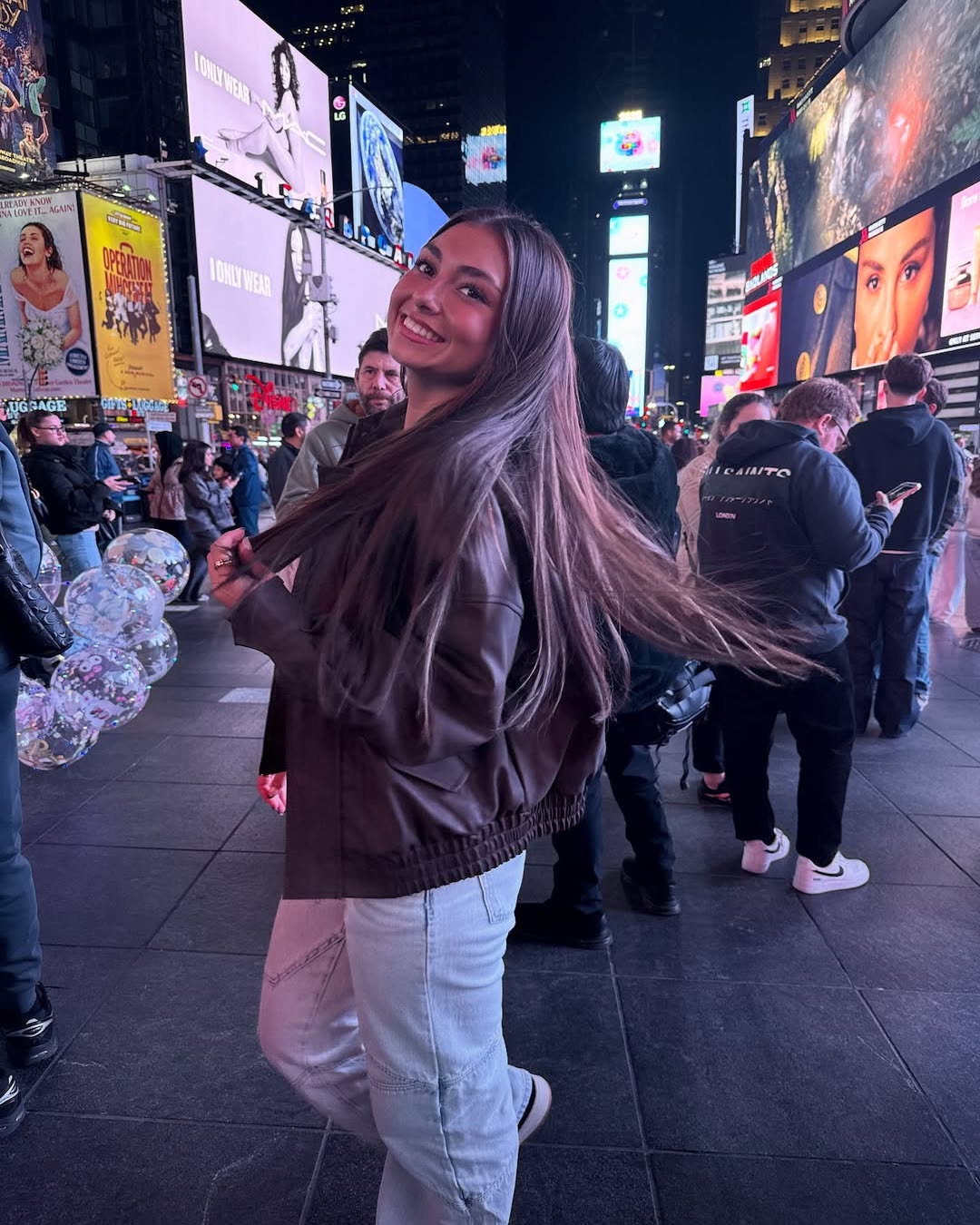 A young woman with long hair smiles and turns toward the camera in Times Square at night, surrounded by people and bright digital billboards. She wears a brown jacket and light jeans, glowing confidently as if she knows all the best beauty hacks.