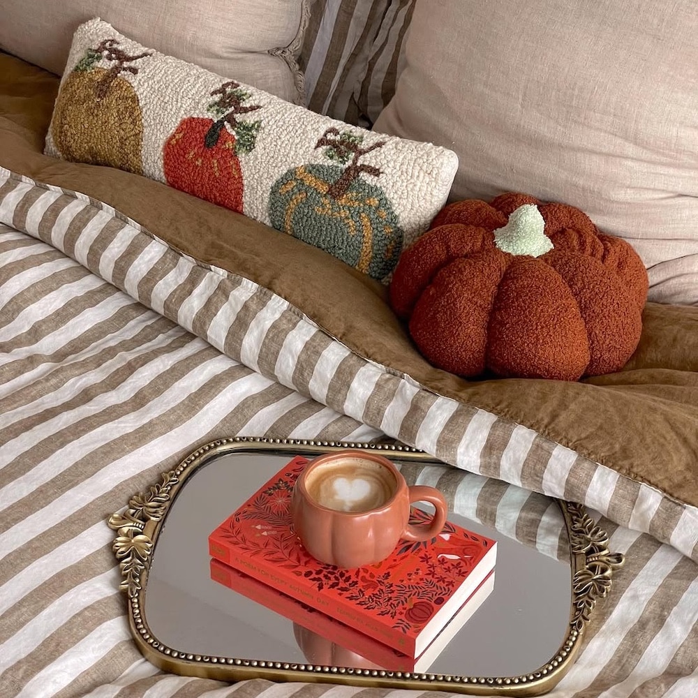 A cozy bed with striped bedding, a pumpkin-themed cushion, and a plush pumpkin pillow. A decorative tray holds a latte in an orange mug and a red book, perfect for inspiring fall bedroom ideas and creating a warm autumnal atmosphere.