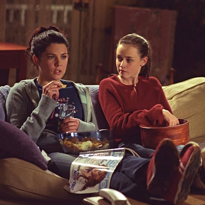 Two women sit on a couch eating snacks; one holds a bowl of chips and the other a wooden bowl. A magazine rests on the lap of the woman in front. They appear to be having a relaxed conversation indoors.