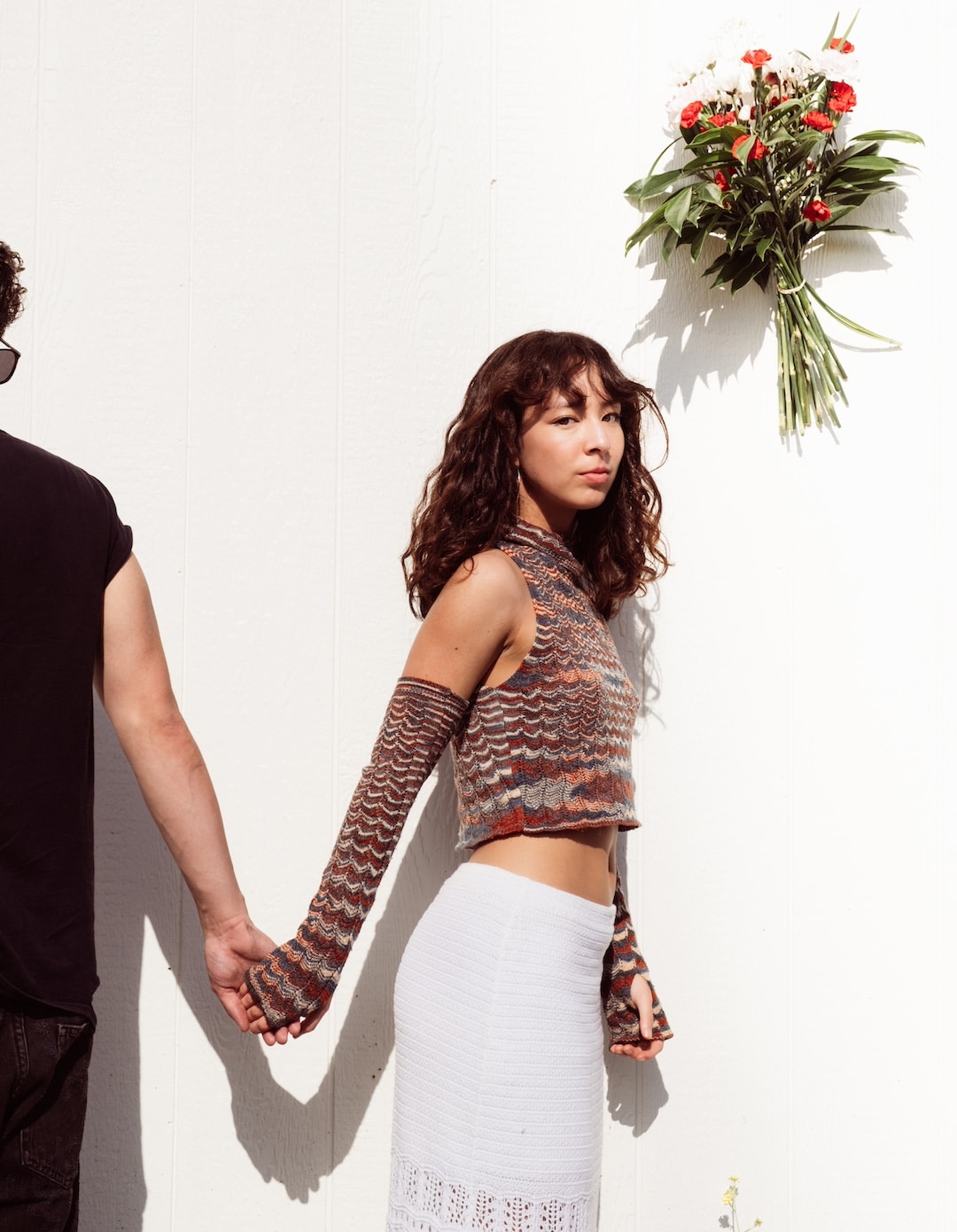A woman with curly hair stands against a white wall, wearing a patterned crop top and white skirt, holding hands with someone who is mostly out of frame. A bouquet of flowers hangs on the wall above her, giving off subtle Frances Anderson fan club vibes.