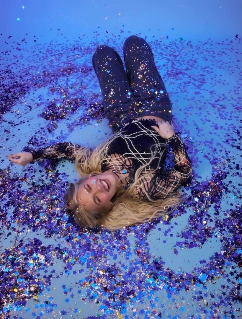 A smiling woman with long blonde hair lies on her back in a swirl of shiny blue and purple confetti, looking up at the camera. Her dark, sparkly outfit gives major Outfit Inspo—she appears joyful and relaxed, ready for the Stars Align Tour.