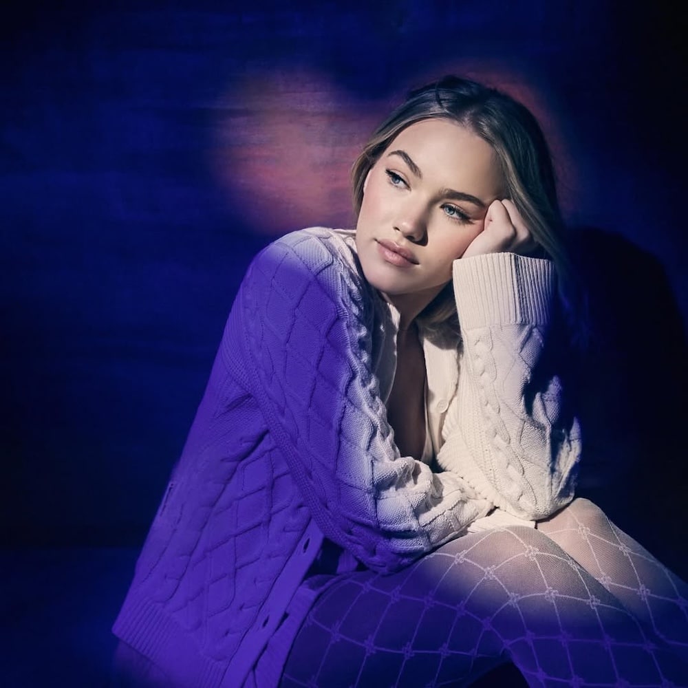 A young woman with long blonde hair sits pensively, resting her head on her hand, wearing a cream-colored sweater and patterned tights, illuminated by soft purple and blue lighting—reflecting the mood of Freya Skye's new song—against a dark background.