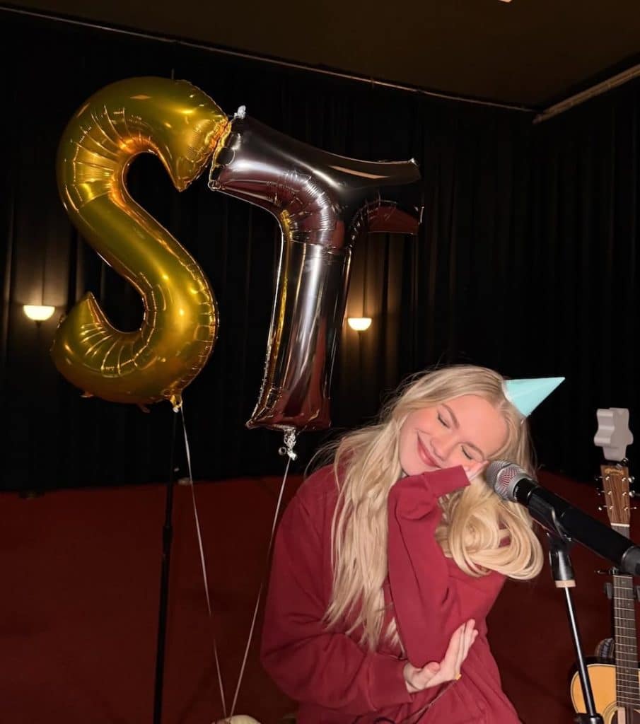A smiling young woman with long blonde hair and a blue party hat sits in front of a microphone, posing happily—perhaps celebrating the release of Freya Skye's new song. Behind her are large metallic gold and silver balloons shaped like the letters S and T.