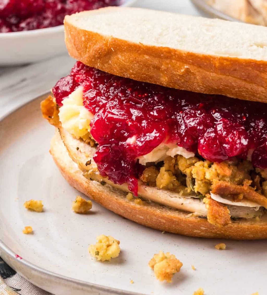 A close-up of a sandwich on white bread filled with stuffing, turkey slices, mashed potatoes, and a generous layer of cranberry sauce—one of the best Friendsgiving sandwiches, with crumbs scattered on the plate.