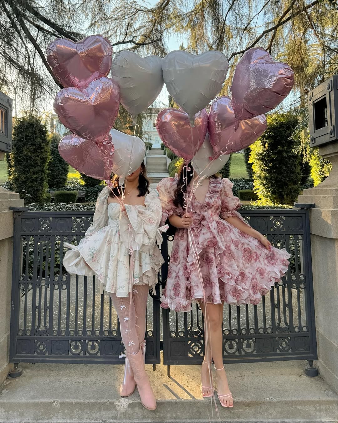 Two women in floral dresses stand side by side, holding pink and silver heart-shaped balloons that cover their faces—perfect inspiration for your next Galentine's Day party. They pose in front of an ornate black gate with trees and greenery behind them.