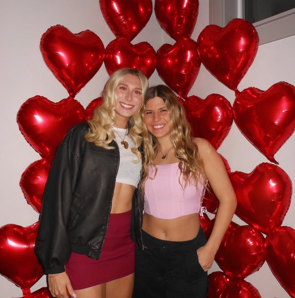 Two women smile in front of a wall adorned with large red heart-shaped balloons at a Galentine's Day party. One wears a black jacket and red skirt; the other, a pink strapless top and black pants.