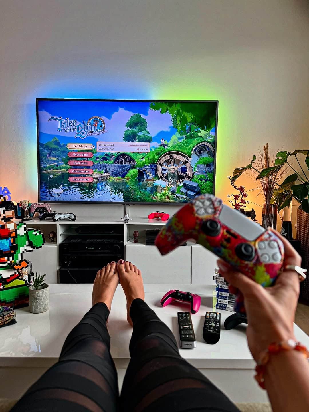 A person with painted nails holds a colorful PlayStation controller while sitting on a couch, enjoying Little Big Planet 3. Gaming consoles, decorations, and plants fill the modern living room—perfect for things to do indoors during freezing weather.