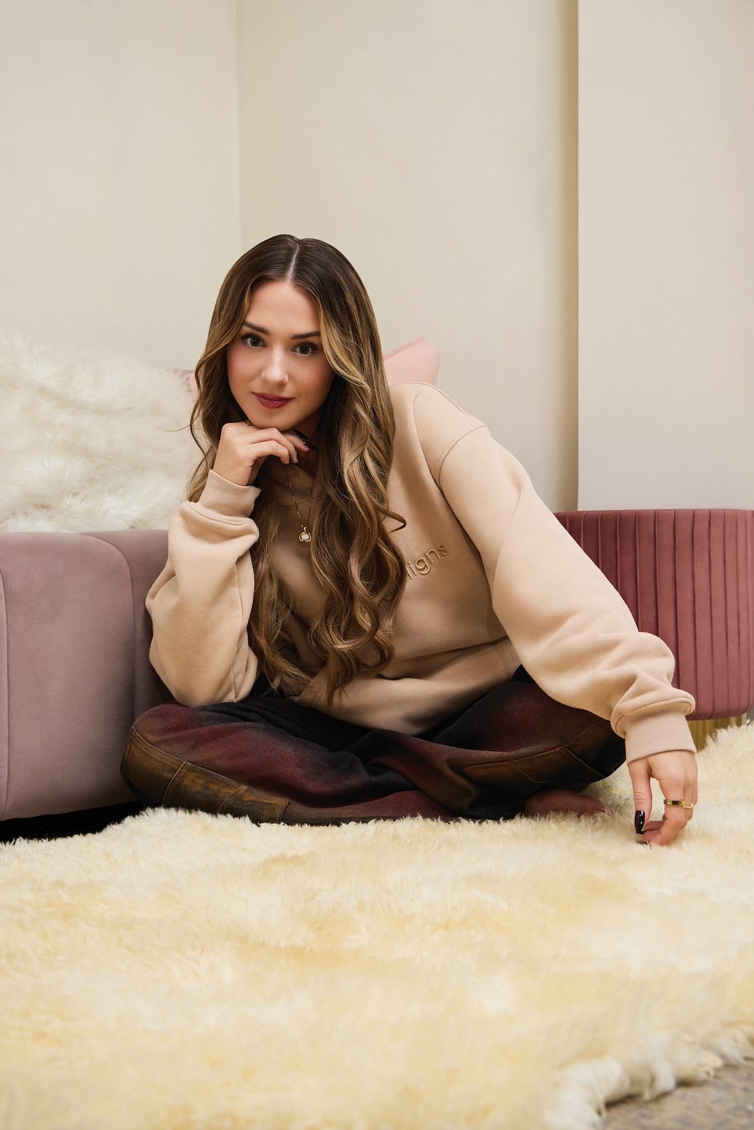 A young woman with long wavy hair sits cross-legged on a cream-colored shaggy rug, leaning on a pink sofa. Gianna Joyce wears a beige sweatshirt and plaid pants, smiling softly at the camera in a cozy indoor setting.