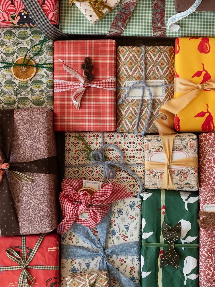 A variety of gift boxes wrapped in colorful, patterned paper and fabric, decorated with ribbons, bows, twine, and natural elements, arranged closely together in a festive display.