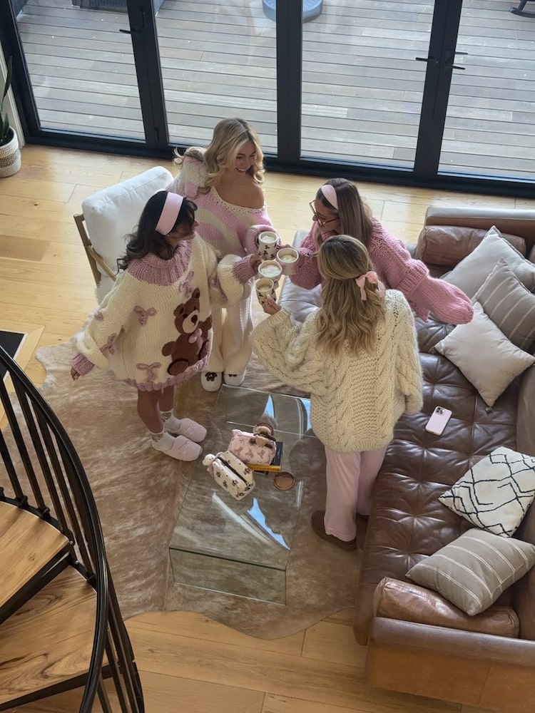 Four women in cozy pastel sweaters and slippers toast with mugs in a living room—one of the best friend hangout ideas and perfect for winter indoor activities when it’s freezing outside. A leather couch and spiral staircase complete the warm scene.