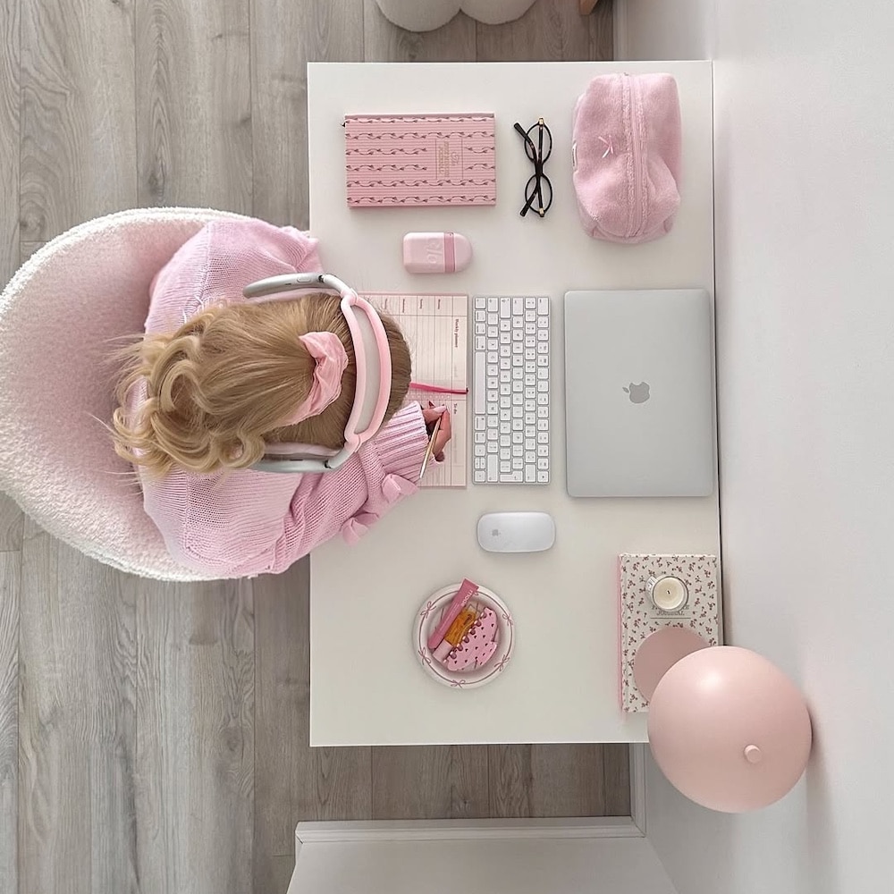 A woman in a pink sweater and headphones sits at a white desk with a laptop, keyboard, mouse, notebook, glasses, pouch, and snacks, all in shades of pink. The workspace is neatly organized and color-coordinated.