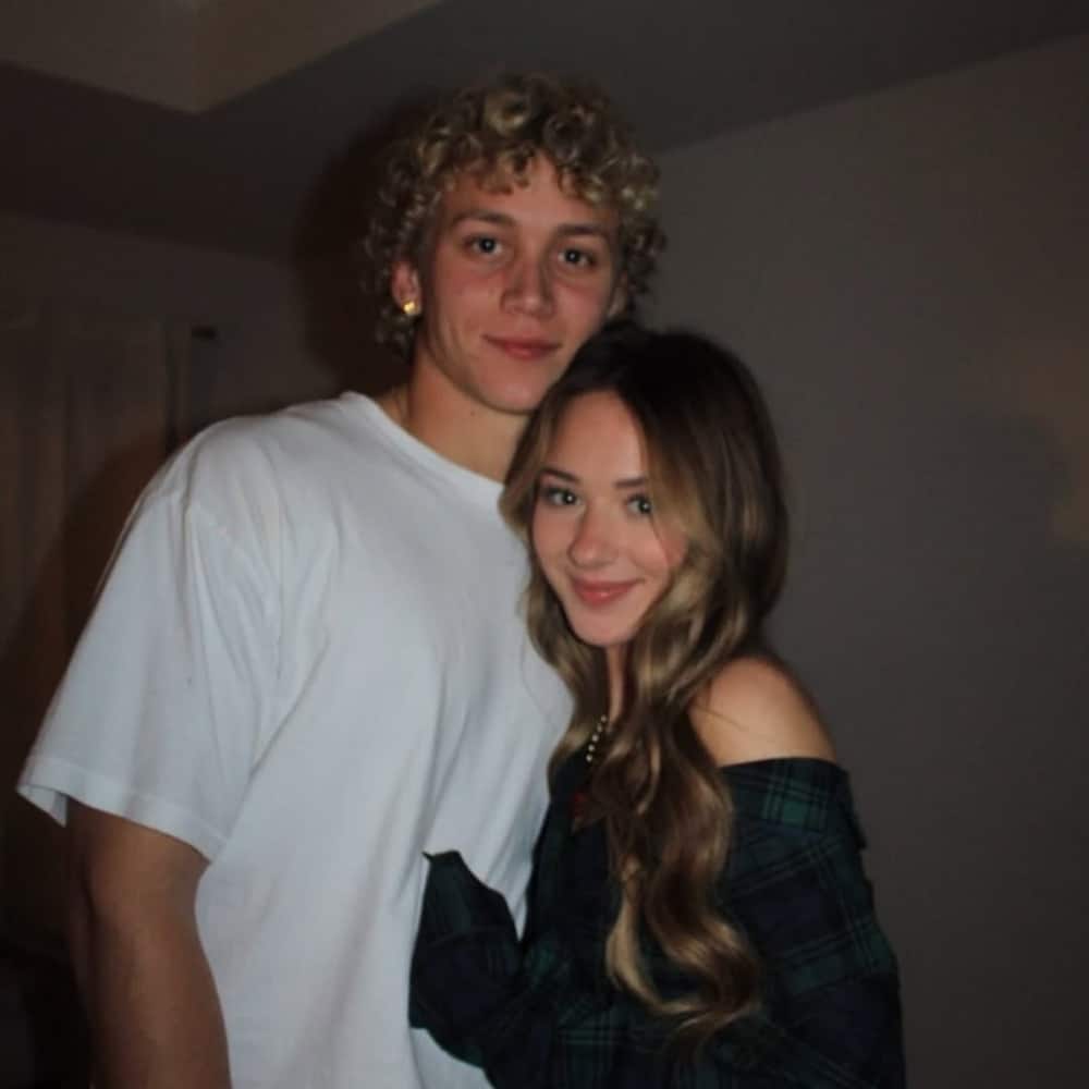 A young couple poses indoors, smiling. This low-key moment features a man with curly blond hair in a white t-shirt and a woman in an off-shoulder green plaid shirt. Their cozy pose is perfect inspiration for sweet date ideas.