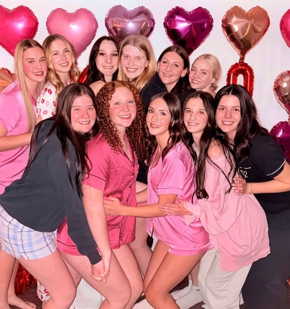 A group of twelve young women in pajamas pose smiling together in front of a wall decorated with pink and red heart-shaped balloons, capturing the perfect Galentine's Day party moment.