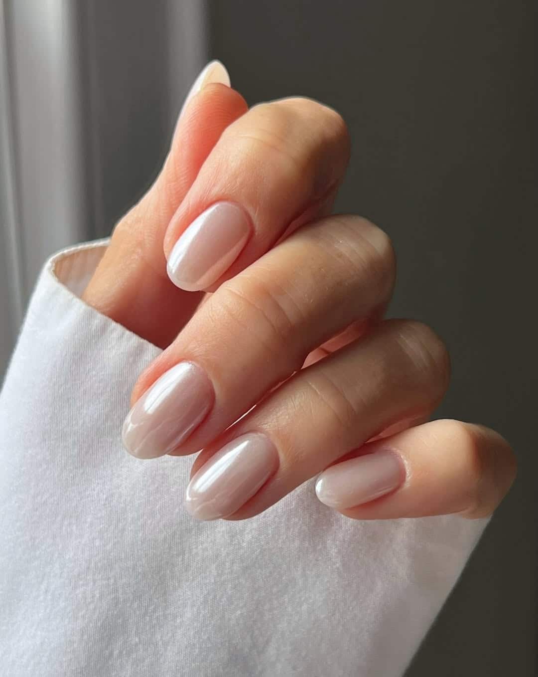 A hand with neatly manicured nails painted in a light, sheer nude color—perfect for 2026 nail designs—partially covered by the sleeve of a white shirt, against a soft, neutral background.