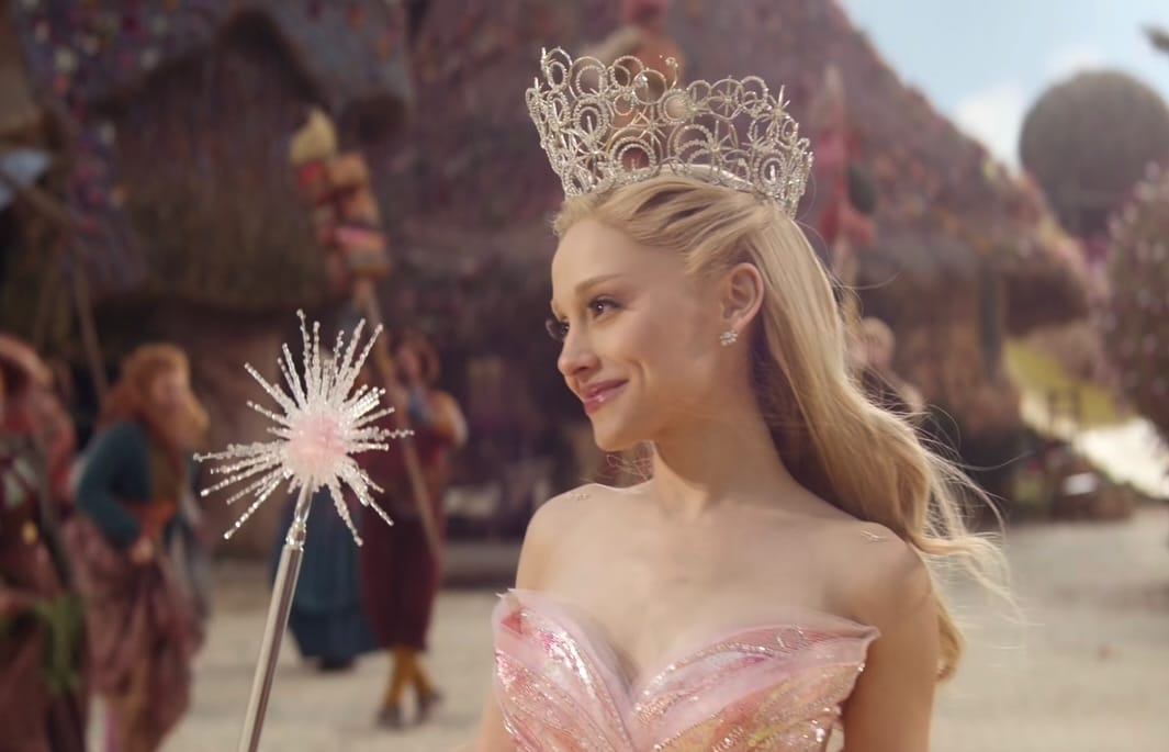 A woman in a sparkling pink dress and elaborate crown smiles while holding a star-shaped wand. She stands outdoors with colorful buildings and people in the background, as if ready to step into theaters for Wicked Part 2’s “For Good.”.